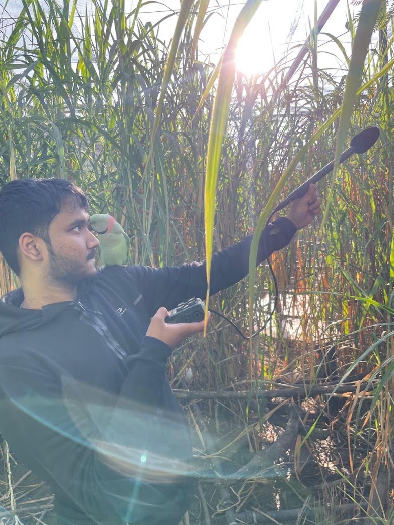 Nakul Wewhare using recording equipment with a parrot perched on his shoulder at a zoo.